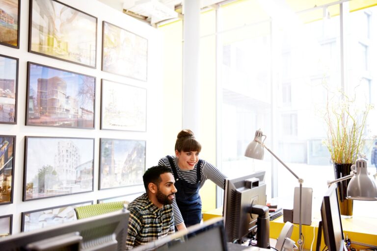 A businessperson standing next to her coworker in an office, looking at the computer together.