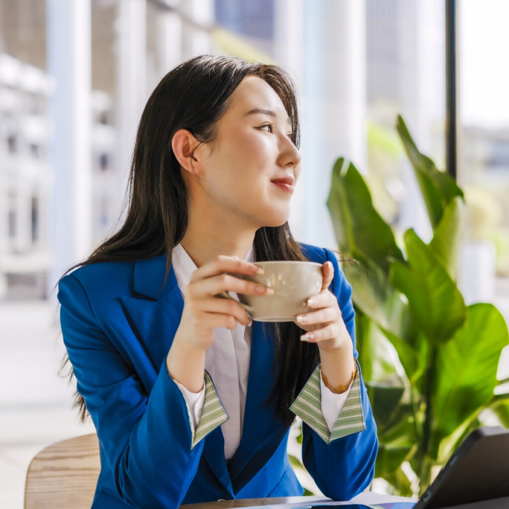 A young Asian businesswoman wearing a blue suit, sat in a bright cafe and drank coffee.