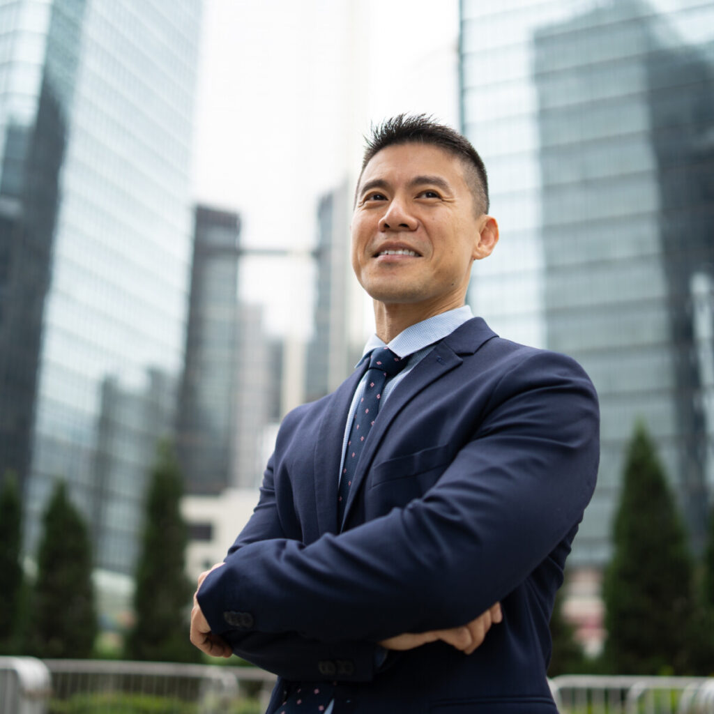 A businessman smiling as he stands with his arms crossed against a background of tall office buildings in a city.