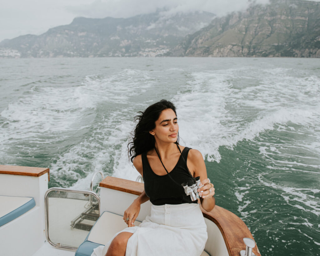 A woman sits on the back of a luxury yacht. As the boat moves along, she looks through a pair of silver binoculars.