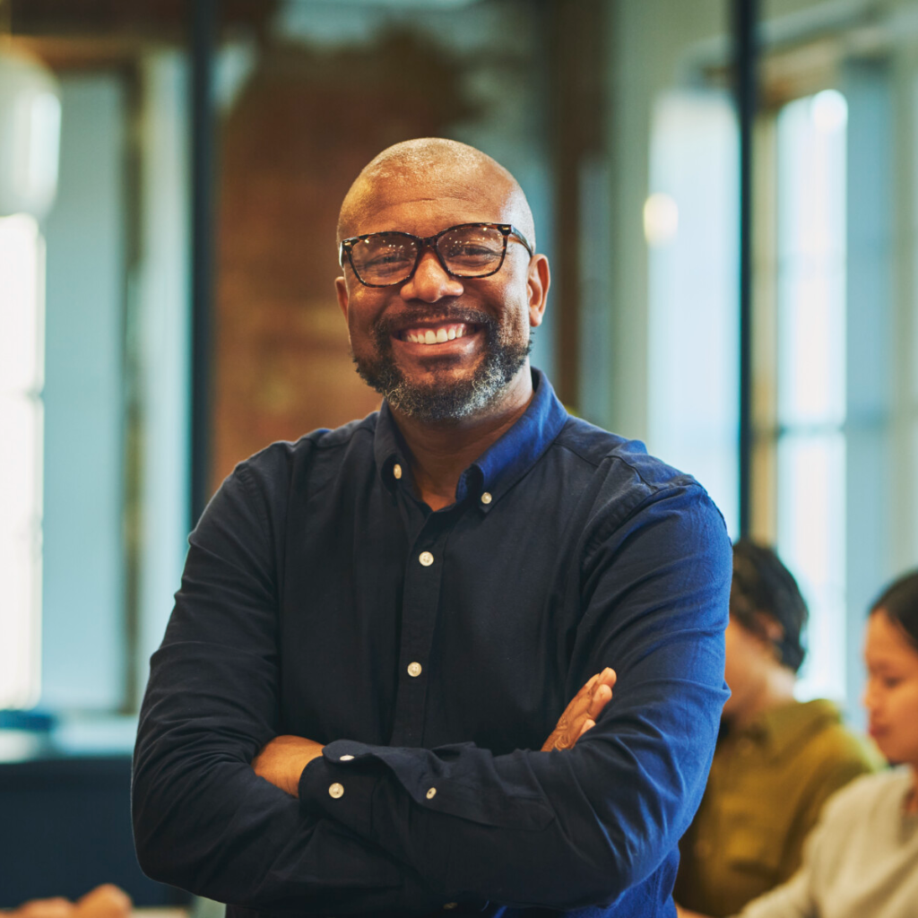 Bearded businessman wearing eyeglasses, crossing arms, and smiling cheerfully in office, colleagues meeting in background.