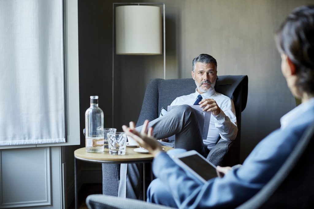 A businessman and a colleague are sitting on chairs during a meeting in a hotel room.