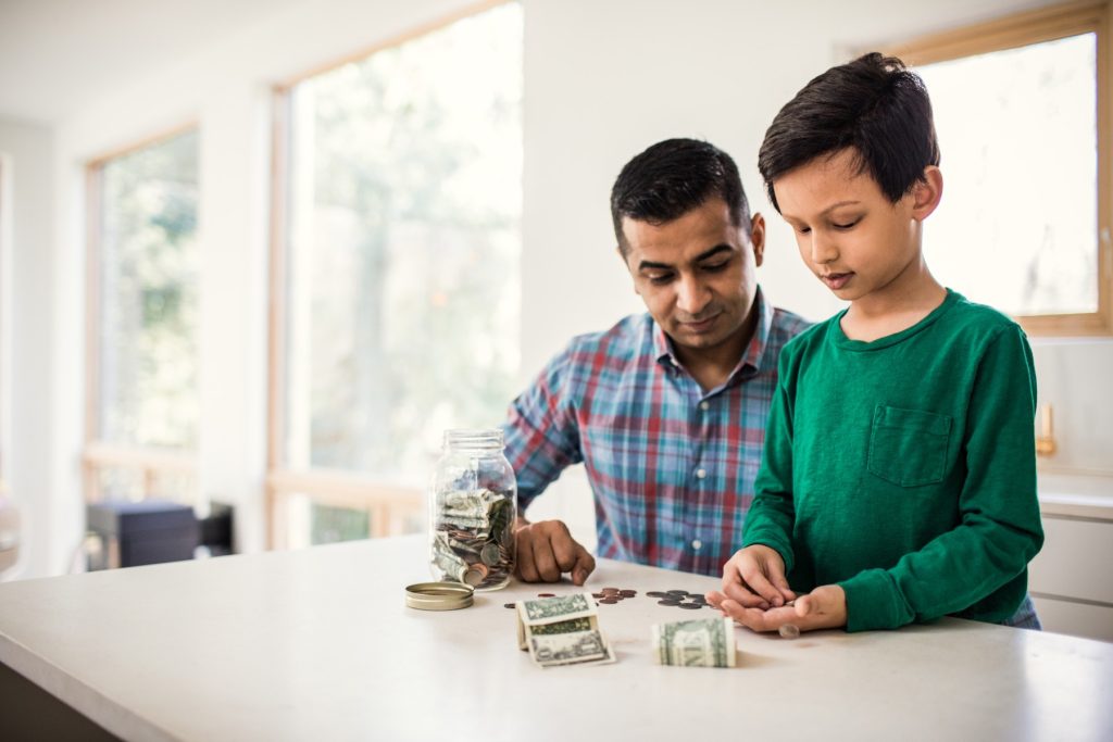 Father and young son sitting at a table, counting and sorting money together at home.