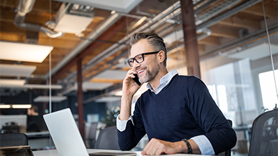 A business professional sitting at a table with a laptop and talking on his mobile phone