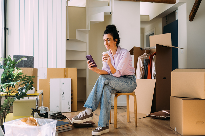 A relaxed female amidst moving boxes in a new apartment, taking a break to use her phone