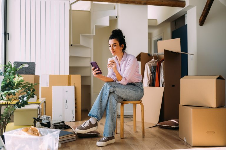 A relaxed female amidst moving boxes in a new apartment, taking a break to use her phone