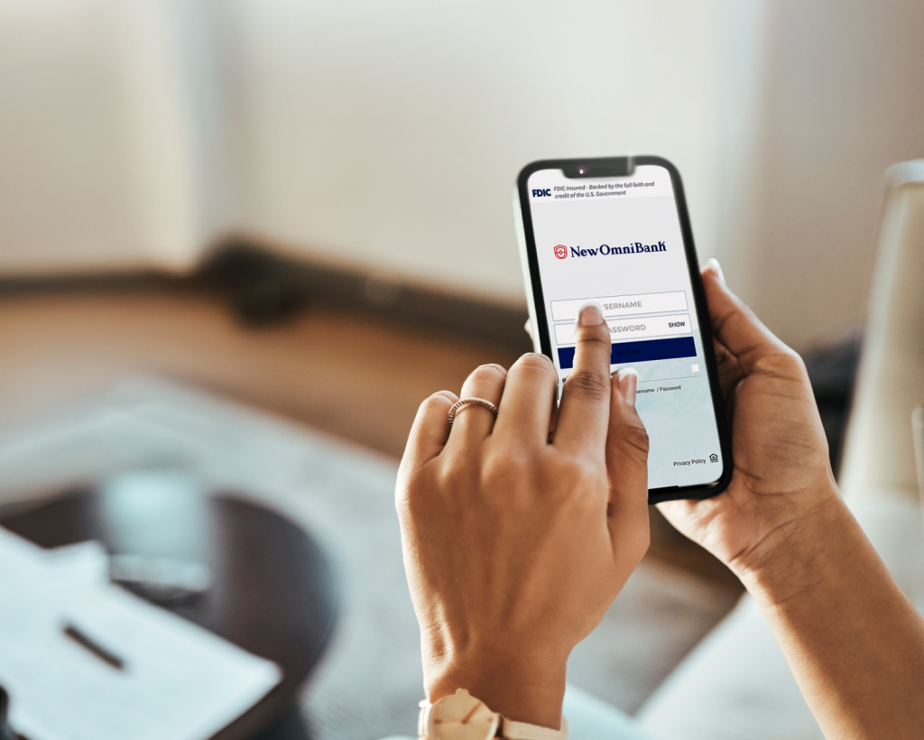 A hand holding a cellphone that shows the login page of New Omni Bank’s banking app.