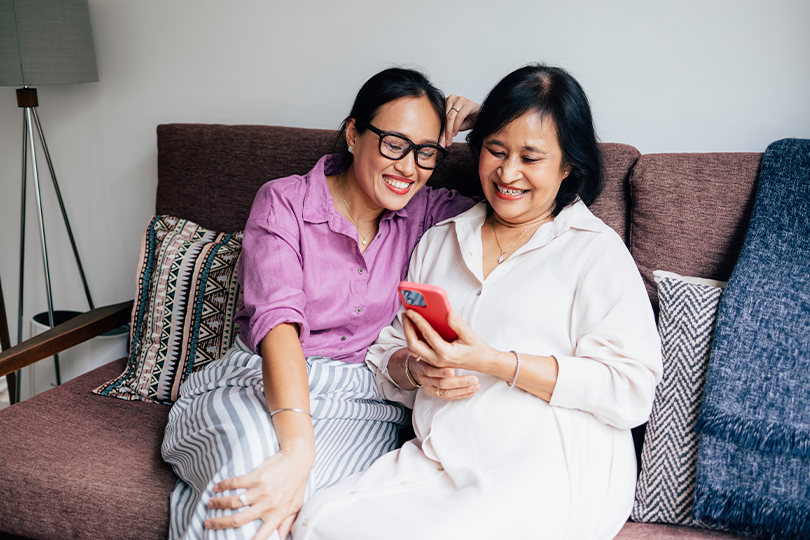 A smiling Asian female reading something using her smartphone while relaxing with her daughter at home.