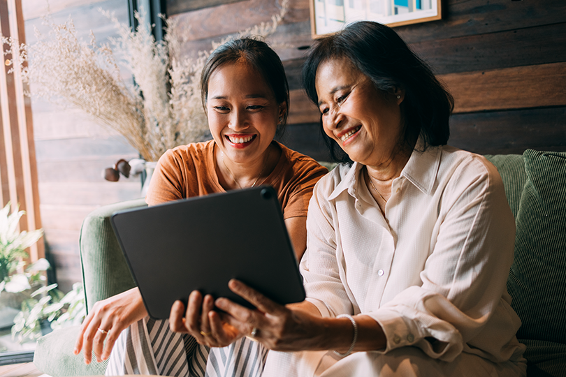 An adult daughter and her mother look at a tablet together at home.