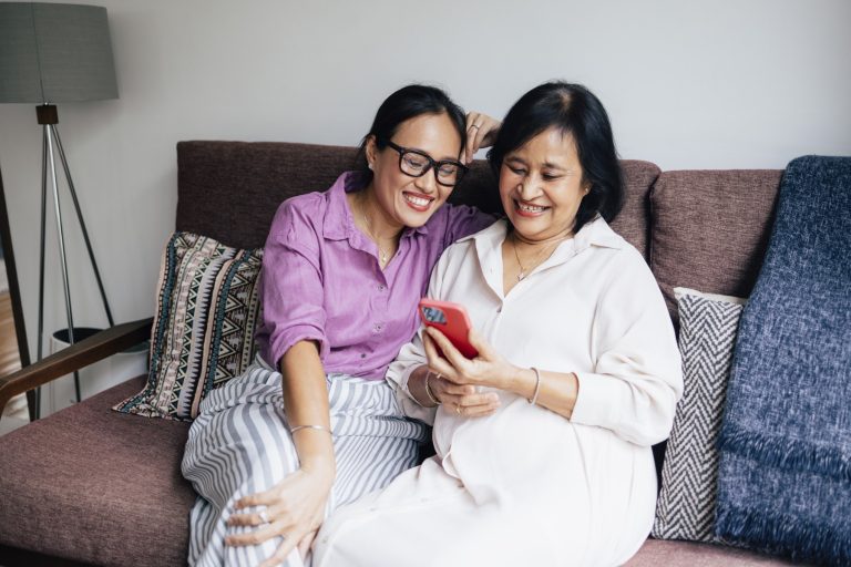 A smiling Asian female reading something using her smartphone while relaxing with her daughter at home.
