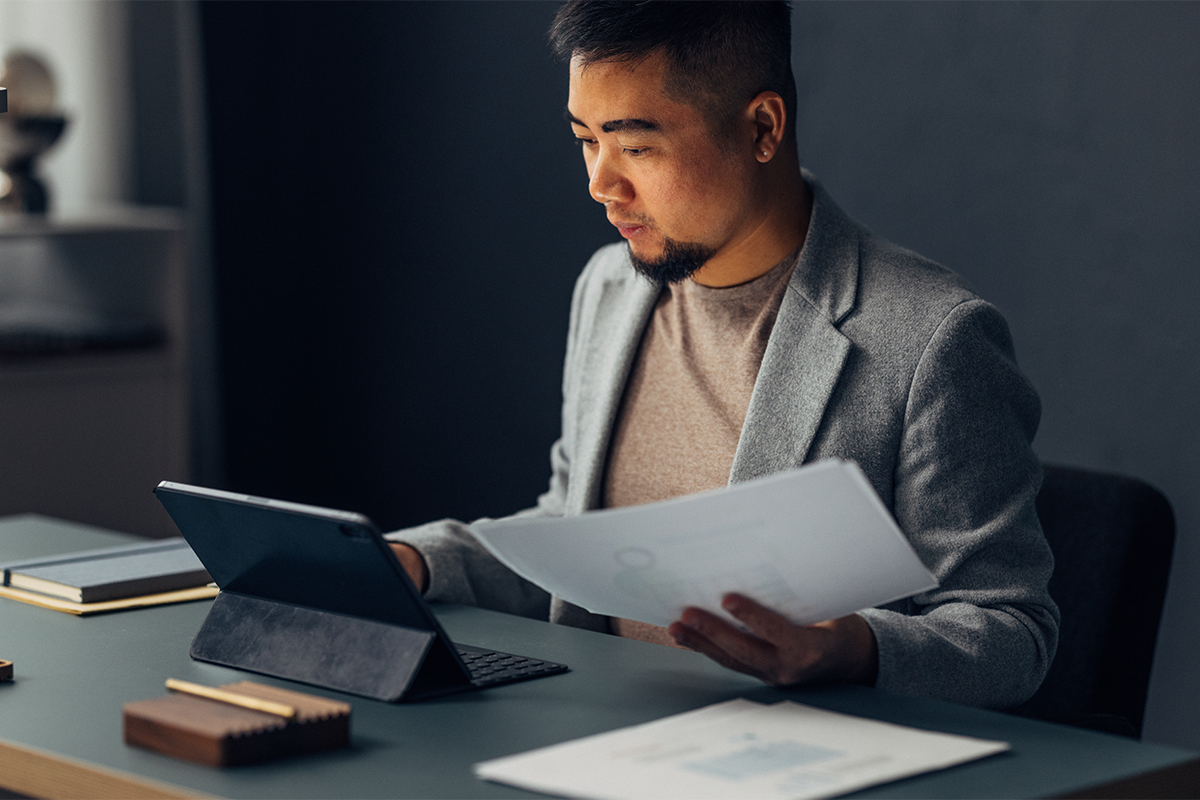 A focused young man analyzing paperwork while working on a tablet in a well-organized home office with mood lighting.