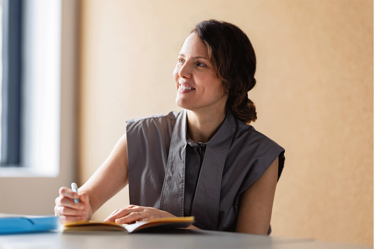 A smiling businesswoman writes in a notebook while looking thoughtfully out of a bright modern office window
