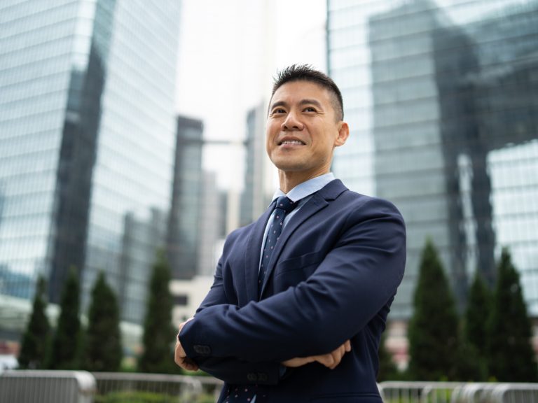 Portrait of a businessman smiling as he looks into the distance, standing with his arms crossed against a background of tall glass covered office buildings in a city.